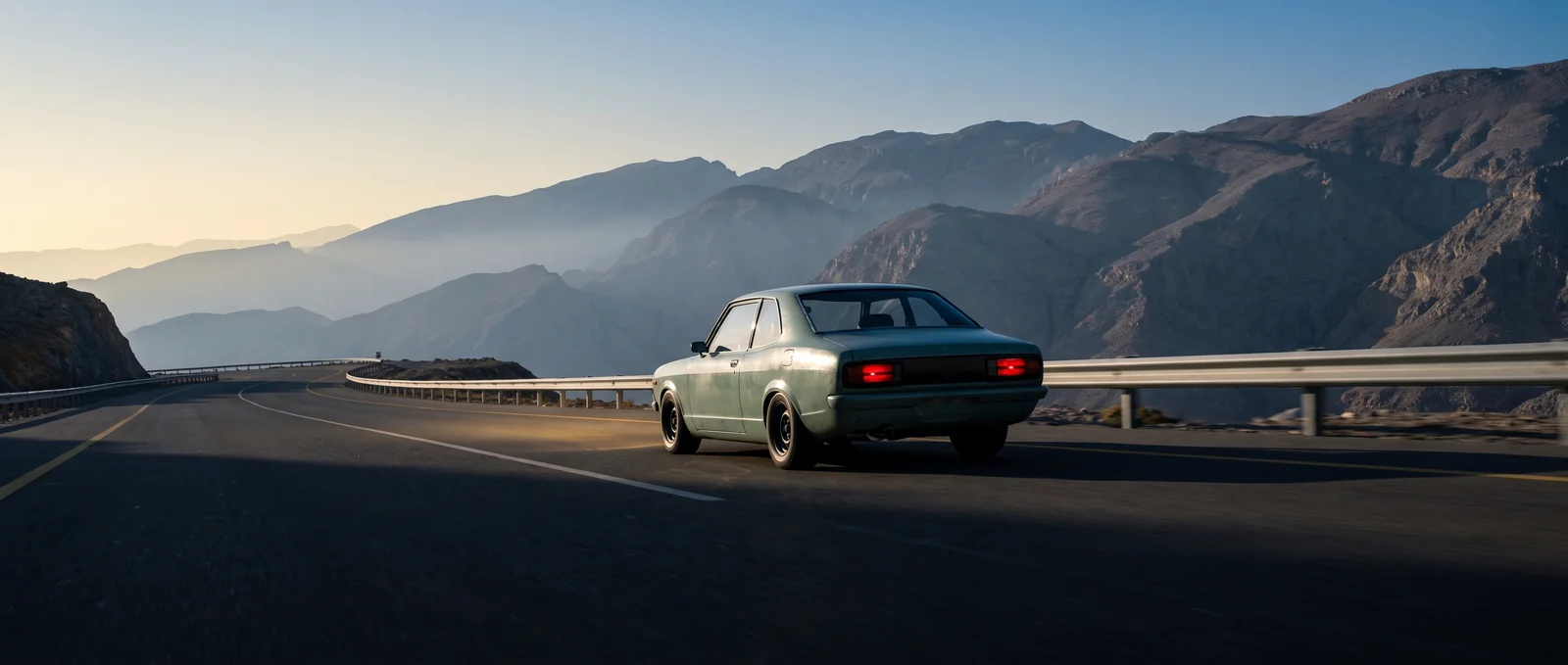 A pale green coupe from the seventies climbs an empty mountain road before sunrise, tail lamps red, ridges fading into blue haze