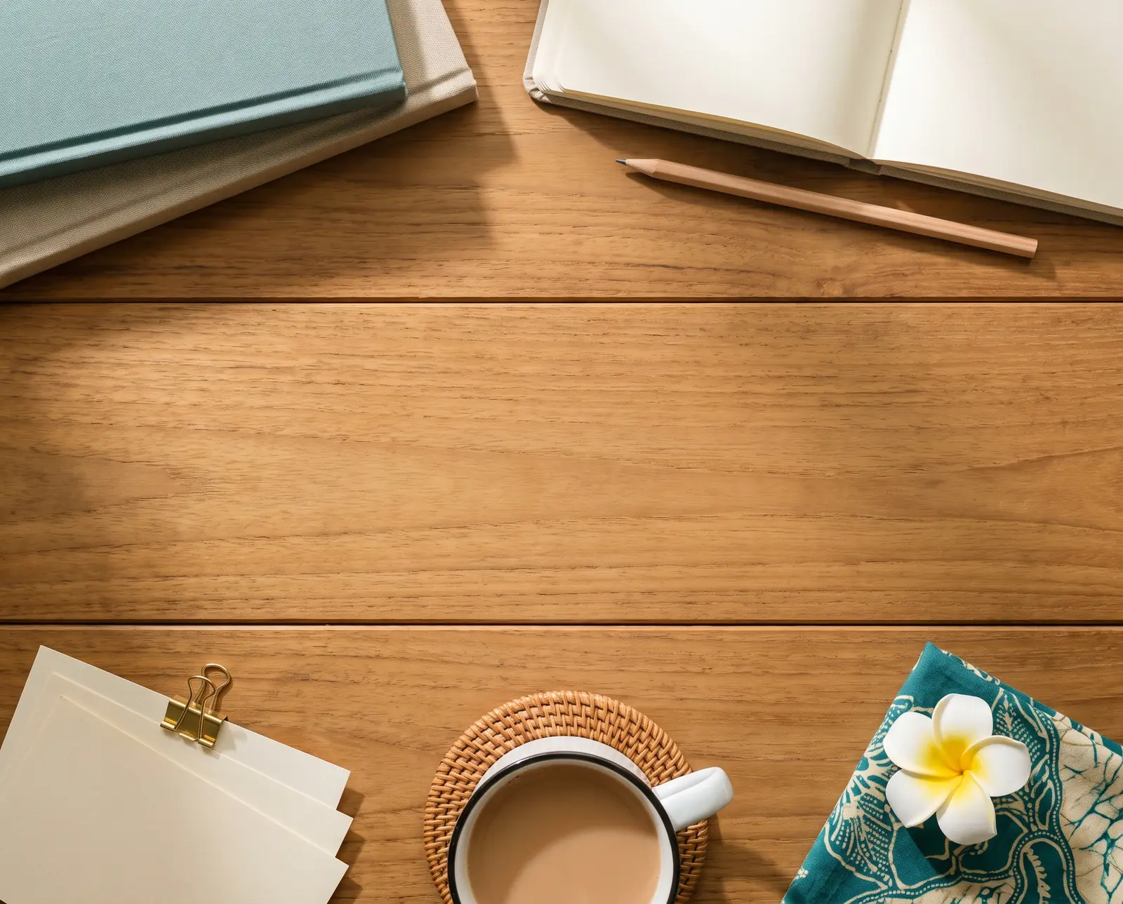A wooden desk from above with notebooks, a pencil, a cup of tea and a frangipani on a batik cloth