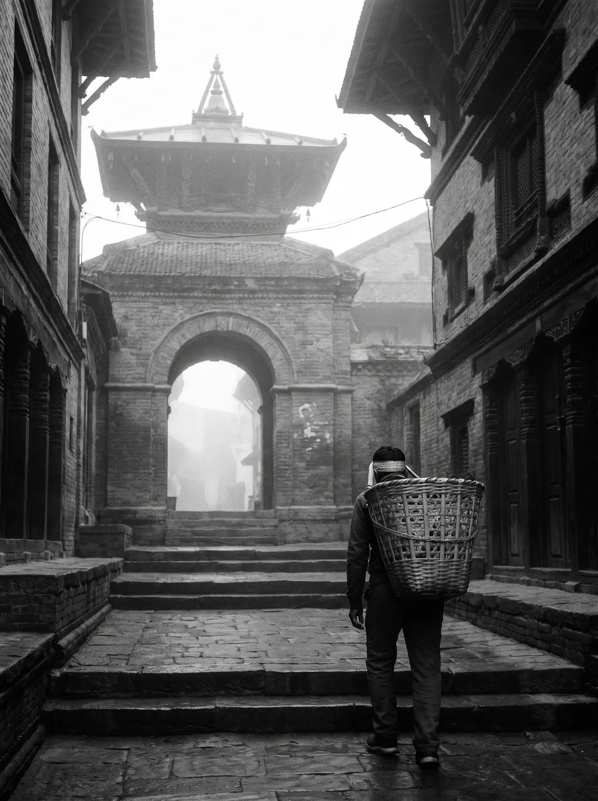 A porter with a woven basket on his back climbs stone steps toward a temple gate in the fog
