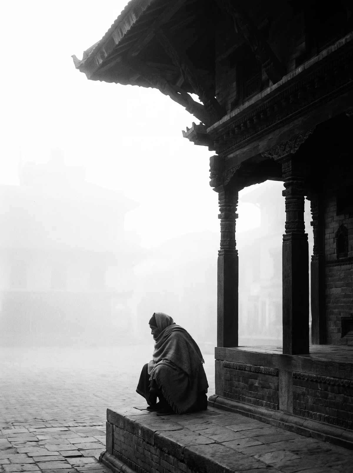 An old man in a shawl sits under a wooden rest house in the fog